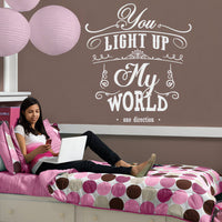 Wall quote sticker with text "You Light Up My World" by One Direction next to a young woman laying on her bed with her laptop.