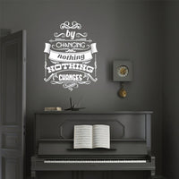 Wall quote sticker with text "By Changing Nothing, Nothing Changes" on a black wall above a piano.