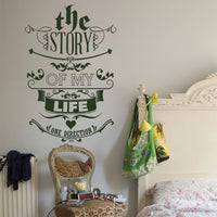 Wall quote sticker with text "The Story Of My Life" by One Direction in a bedroom near a chair and a bed.
