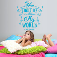 Wall quote sticker with text "You Light Up My World" by One Direction above a little girl resting on some cushions.